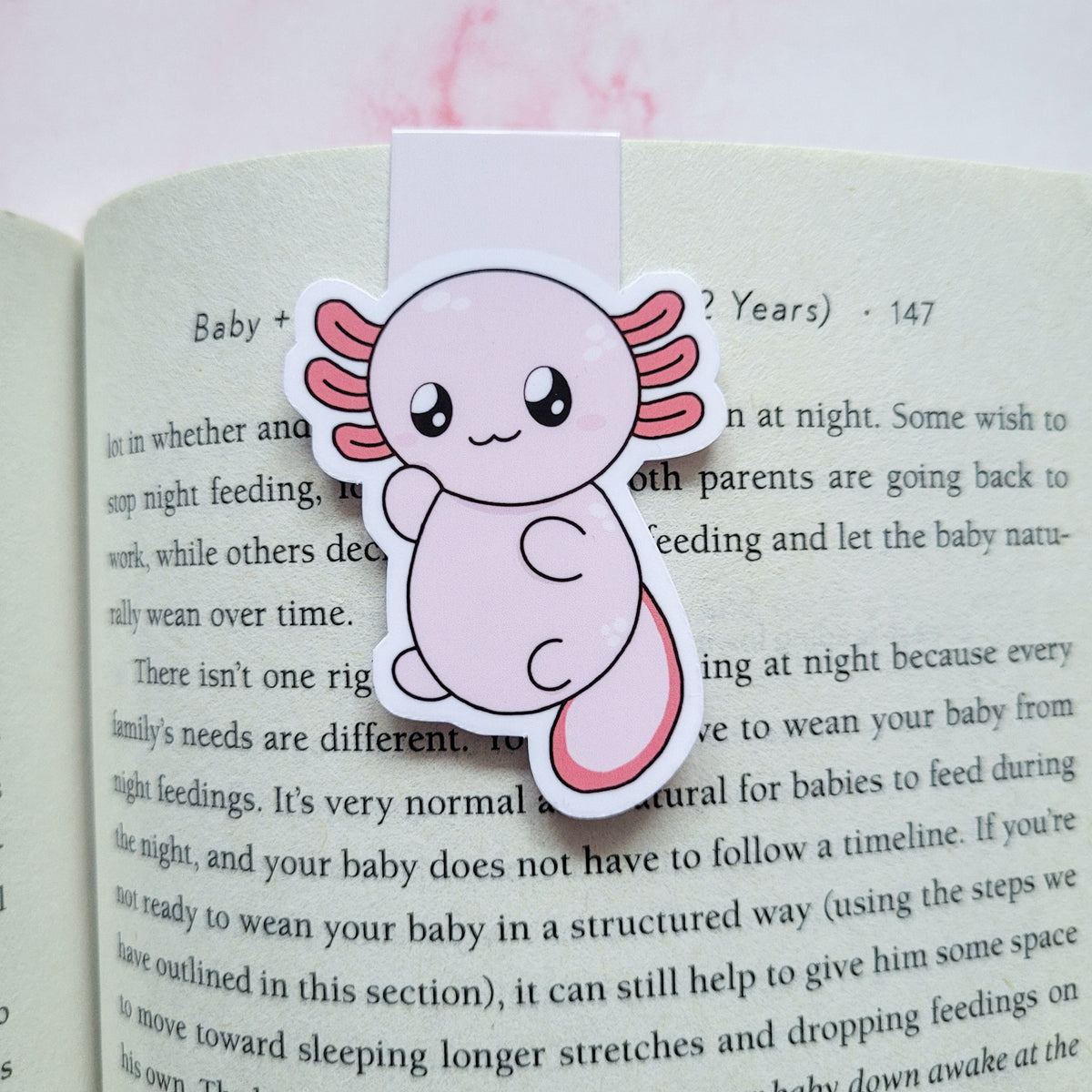 A cute die-cut sticker of a smiling pink axolotl fish, designed as a book bookmark, shown clipped onto an open book page.