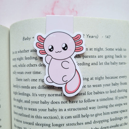 A cute die-cut sticker of a smiling pink axolotl fish, designed as a book bookmark, shown clipped onto an open book page.